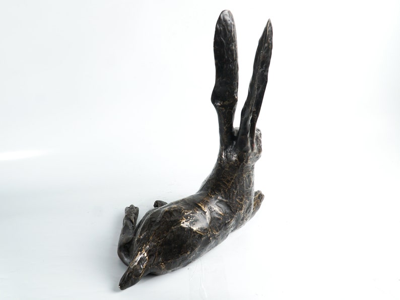 K&ouml;nnte beinhalten: Eine Bronzeskulptur eines liegenden Hasen mit erhobenen Ohren. Die Skulptur hat eine strukturierte Oberfl&auml;che und eine dunkelbraune Farbe.