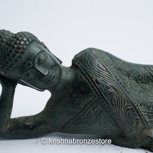 May include: A green bronze statue of a reclining Buddha with intricate details on the clothing. The Buddha has closed eyes and is resting the head on the left hand.