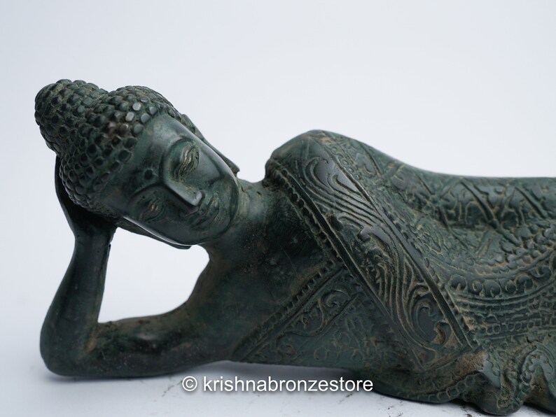 May include: A green bronze statue of a reclining Buddha with intricate details on the clothing. The Buddha has closed eyes and is resting the head on the left hand.