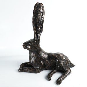 K&ouml;nnte beinhalten: Eine Bronzeskulptur eines liegenden Hasen mit erhobenen Ohren. Die Skulptur ist detailliert mit strukturiertem Fell und hat eine dunkelbraune Patina.
