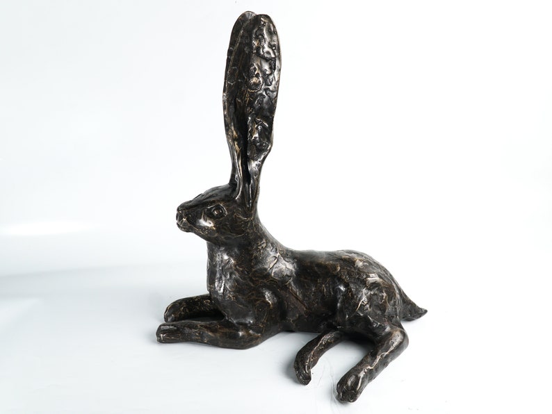 K&ouml;nnte beinhalten: Eine Bronzeskulptur eines liegenden Hasen mit erhobenen Ohren. Die Skulptur ist detailliert mit strukturiertem Fell und hat eine dunkelbraune Patina.