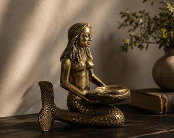 May include: A metallic gold-toned mermaid figurine in a seated position, featuring a textured fish tail and long hair. The mermaid holds a shallow, shell-shaped bowl in both hands, with a weathered finish and visible sculpted details throughout.