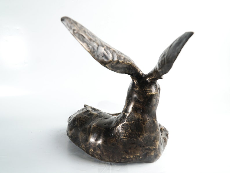 K&ouml;nnte beinhalten: Eine Bronzeskulptur eines Hasen mit erhobenen Ohren, der Hase liegt mit dem Kopf zur Seite gedreht.