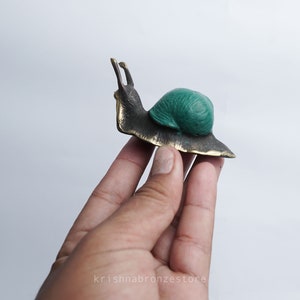 May include: A small bronze snail figurine with a green shell. The snail is sitting on a hand.