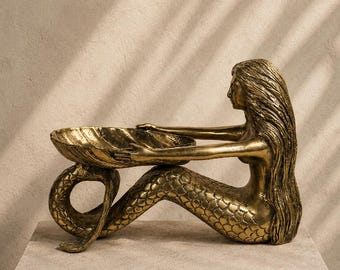 May include: A metallic gold-toned sculpture of a mermaid with long flowing hair, depicted in a seated position with her tail curled. She holds a large, open seashell-shaped bowl in her outstretched arms, featuring detailed scale textures.