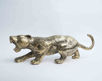 May include: A gold-colored metal figurine of a snarling panther or jaguar. The figurine is posed in a crouching position, with its head turned to the side and its mouth open in a snarl. The figurine is detailed with textured fur and sharp claws.