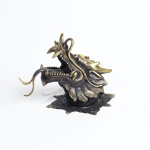 May include: A gold and brown dragon head sculpture with an open mouth and a long, curled tongue. The dragon is mounted on a base with a starburst design.