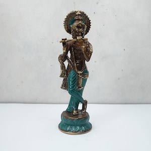 Puede incluir: Una estatua de bronce de Krishna tocando la flauta, con un esquema de colores verde y dorado. La estatua está de pie sobre una base de loto.