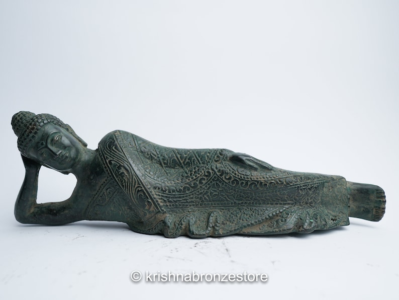 May include: A green bronze statue of a reclining Buddha with intricate details on the clothing. The statue is lying on its side with its head resting on its hand.