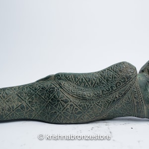 May include: A green bronze reclining Buddha statue with intricate carvings on the robe and head. The statue is lying on its side with its head resting on its hand.