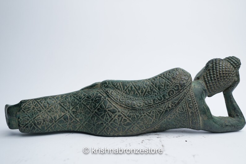 May include: A green bronze reclining Buddha statue with intricate carvings on the robe and head. The statue is lying on its side with its head resting on its hand.