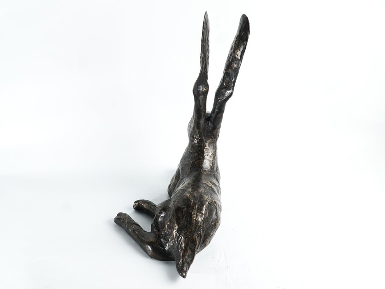K&ouml;nnte beinhalten: Eine Bronzeskulptur eines Hasen mit hoch erhobenen Ohren. Der Hase liegt auf der Seite mit dem Kopf zur Seite gedreht. Die Skulptur ist detailliert und realistisch.