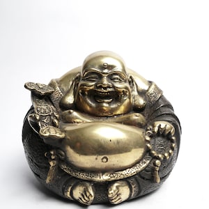 May include: A brass-colored laughing Buddha statue. The figure is seated with a large belly, holding a bag and prayer beads. The statue has a detailed, textured appearance and is set against a white background.