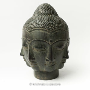 May include: A dark green stone sculpture of a four-faced Buddha head with closed eyes. The sculpture is on a white background.