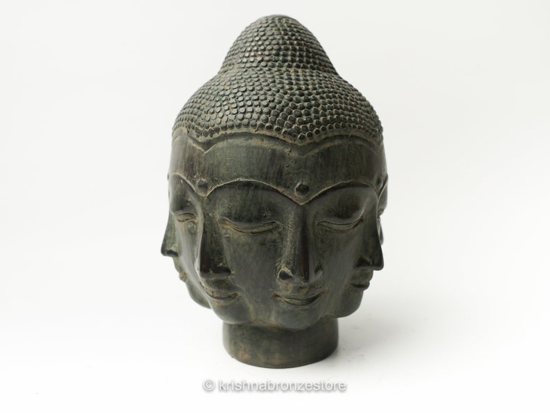 May include: A dark green stone sculpture of a four-faced Buddha head with closed eyes. The sculpture is on a white background.