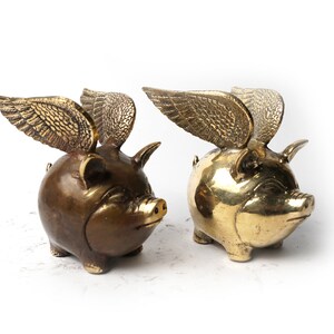 Puede incluir: Dos figuritas de cerdo de latón con alas. Un cerdo es dorado y el otro es marrón.