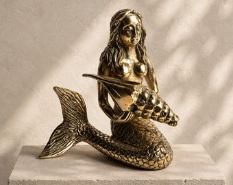 May include: A metallic gold-toned mermaid figurine featuring a textured fish tail and long hair. The mermaid is depicted in a seated position, holding a large, spiraled conch shell in both hands against the chest. The surface has a polished finish.