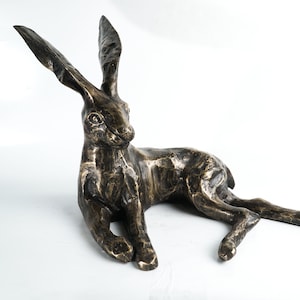 K&ouml;nnte beinhalten: Eine Bronzeskulptur eines liegenden Hasen mit aufgestellten Ohren. Die Skulptur ist detailliert und realistisch, mit einer strukturierten Oberfl&auml;che.