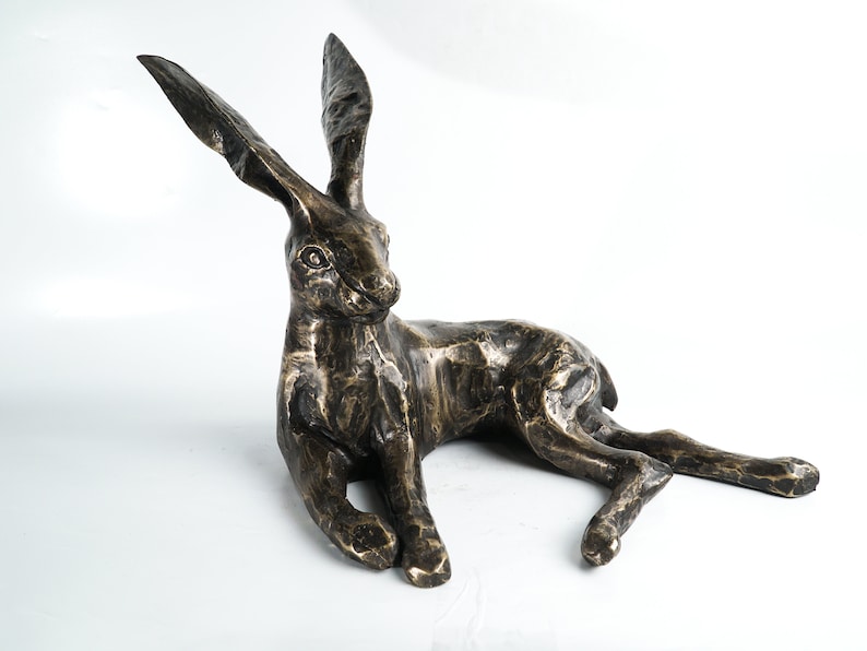K&ouml;nnte beinhalten: Eine Bronzeskulptur eines liegenden Hasen mit aufgestellten Ohren. Die Skulptur ist detailliert und realistisch, mit einer strukturierten Oberfl&auml;che.