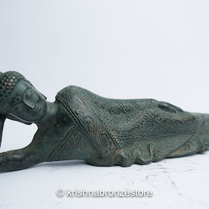 May include: A green bronze statue of a reclining Buddha with intricate details on the robe. The Buddha is lying on its side with its head resting on its hand.