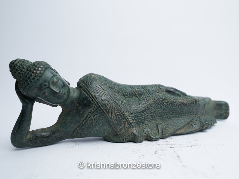 May include: A green bronze statue of a reclining Buddha with intricate details on the robe. The Buddha is lying on its side with its head resting on its hand.