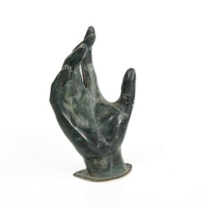 Könnte beinhalten: Eine bronzefarbene Skulptur einer Hand, die Finger leicht gekrümmt nach oben. Die Hand ist auf einem kleinen, rechteckigen Sockel befestigt. Die Skulptur hat eine verwitterte Erscheinung, mit einer grün-braunen Patina.
