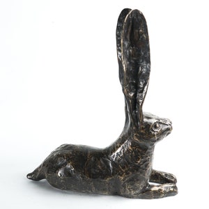 K&ouml;nnte beinhalten: Eine Bronzeskulptur eines liegenden Hasen mit erhobenen Ohren. Die Skulptur ist dunkelbraun und hat eine strukturierte Oberfl&auml;che.