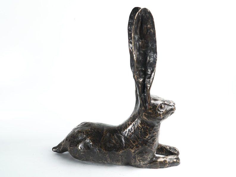 K&ouml;nnte beinhalten: Eine Bronzeskulptur eines liegenden Hasen mit erhobenen Ohren. Die Skulptur ist dunkelbraun und hat eine strukturierte Oberfl&auml;che.