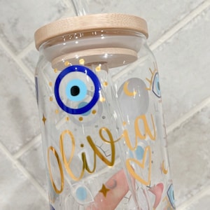 May include: Clear glass tumbler with a bamboo lid and a glass straw. The tumbler is decorated with gold lettering that spells "Olivia", blue evil eye symbols, and gold stars. The tumbler is designed for cold beverages.