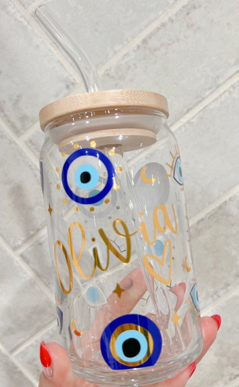 May include: Clear glass tumbler with a bamboo lid and a glass straw. The tumbler is decorated with gold lettering that spells "Olivia", blue evil eye symbols, and gold stars. The tumbler is designed for cold beverages.