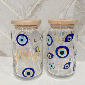 May include: Two clear glass drinking jars with bamboo lids and glass straws. The jars are decorated with blue and gold evil eye designs and the name "Olivia" in gold script. The jars are on a white surface.