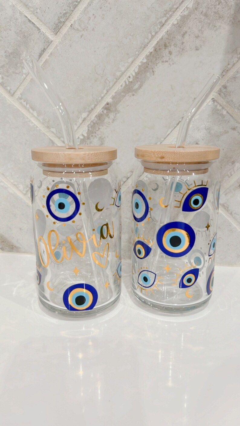 May include: Two clear glass drinking jars with bamboo lids and glass straws. The jars are decorated with blue and gold evil eye designs and the name "Olivia" in gold script. The jars are on a white surface.