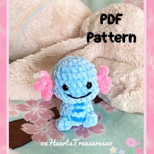 May include: A blue crocheted axolotl plush toy with pink ears, sitting on a white fluffy blanket. The text "PDF Pattern" is written in black on a white background. The text "oxHeartsTreasuresxo" is written in black on a white background.