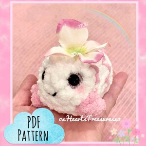May include: A handmade, plush, white and pink creature with large black eyes and a flower on its head. The creature has pink accents and is held in a hand. The image includes the text "PDF PATTERN" in a blue cloud and the shop name.
