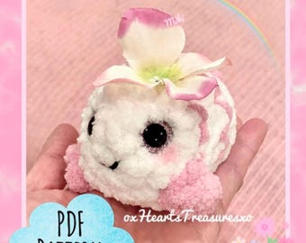 May include: A handmade, plush, white and pink creature with large black eyes and a flower on its head. The creature has pink accents and is held in a hand. The image includes the text "PDF PATTERN" in a blue cloud and the shop name.