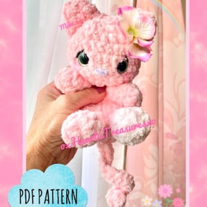 Puede incluir: Un juguete de ganchillo amigurumi de gato rosa con una flor en la cabeza. El juguete está siendo sostenido por una mano. El texto "PDF PATTERN" es visible en la imagen.