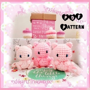 May include: Three pink crocheted amigurumi plushies in the shape of a cat. The plushies are sitting on a white surface with a pink and white floral pattern. The text "enjoy the little things" is printed on the surface. The text "xHearts Treasuresxo" is printed on the bottom of the image.