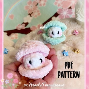 Pode incluir: Dois peluches de concha de amêijoa de crochê, um rosa e outro verde claro, com uma pérola branca de crochê no interior. Os peluches estão sentados em uma superfície branca fofa com um arco-íris e flores rosa no fundo. O texto "PDF PATTERN" está escrito em preto sobre um fundo branco.