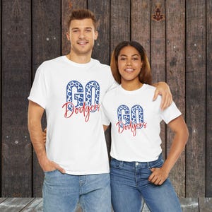 May include: Two people wearing white t-shirts with the text "GO Dodgers" in blue leopard print and red script. The shirts are paired with blue jeans. The background is a dark wood panel.
