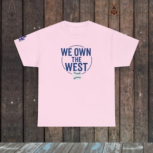 May include: A light pink t-shirt with the text "WE OWN THE WEST" in navy blue, encircling a baseball graphic. The shirt is displayed on a wooden surface.