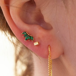 Puede incluir: Un par de pendientes de oro con piedras preciosas verdes. Un pendiente es de forma cuadrada pequeña, y el otro es un racimo de piedras preciosas verdes en forma de hoja.