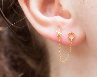 Op de afbeelding: Gouden kettingoorbellen met een klein kruis en een cirkelvormige hanger. De ketting verbindt de twee hangers en creëert een unieke en stijlvolle look.