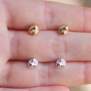 May include: Four small earrings, two gold and two silver, in the shape of a rough, irregular rock. The earrings are shown on a person's hand.