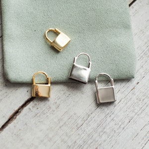 May include: Four small lock charms, two gold and two silver, on a light wood surface. The charms are all the same size and shape, and they have a simple, minimalist design.