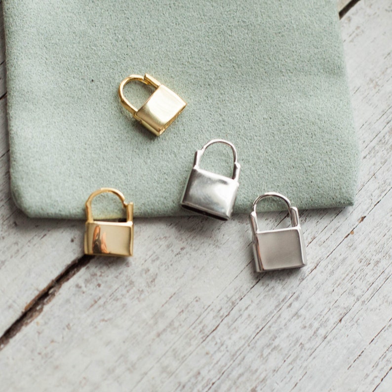 May include: Four small lock charms, two gold and two silver, on a light wood surface. The charms are all the same size and shape, and they have a simple, minimalist design.