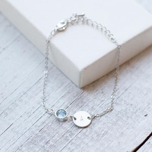 May include: A silver chain bracelet with a small blue gemstone and a round charm engraved with the letter "A".