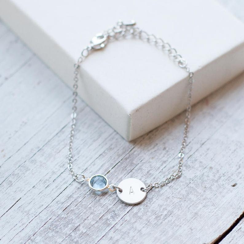 May include: A silver chain bracelet with a small blue gemstone and a round charm engraved with the letter "A".
