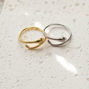 May include: Two snake-shaped rings, one gold and one silver, with green gemstones for eyes. The rings are on a white textured surface.