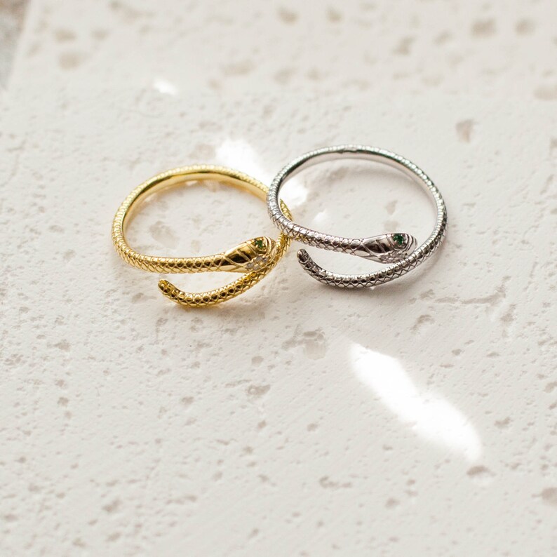 May include: Two snake-shaped rings, one gold and one silver, with green gemstones for eyes. The rings are on a white textured surface.