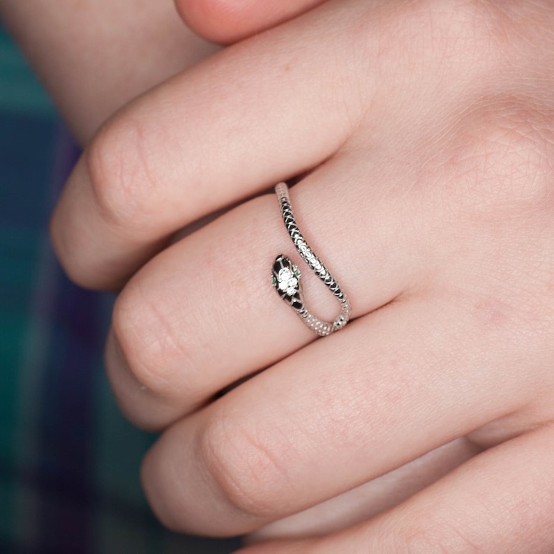 May include: A silver snake ring with a clear stone on the head. The ring is wrapped around a finger.